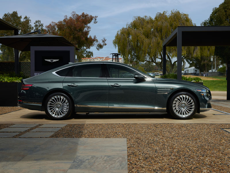 2026 Genesis G80 luxury sedan in green, side profile, parked outdoors on a clear day with trees in the background.