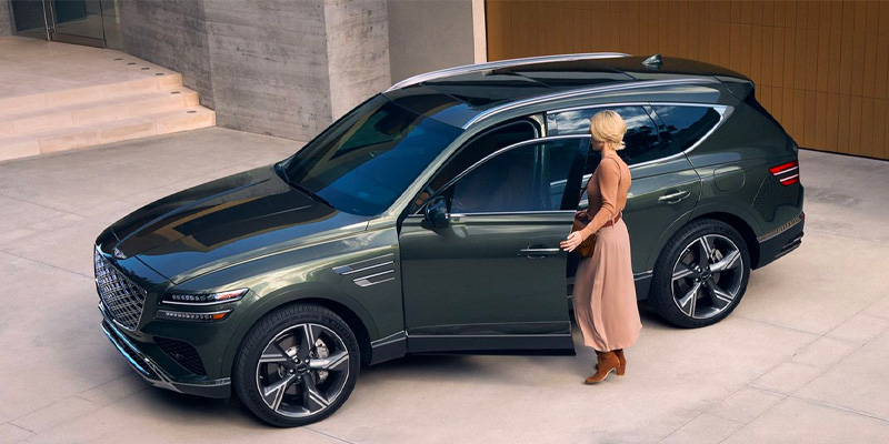 Woman opening the driver door of a green Genesis GV80 luxury SUV parked outside a modern upscale building.
