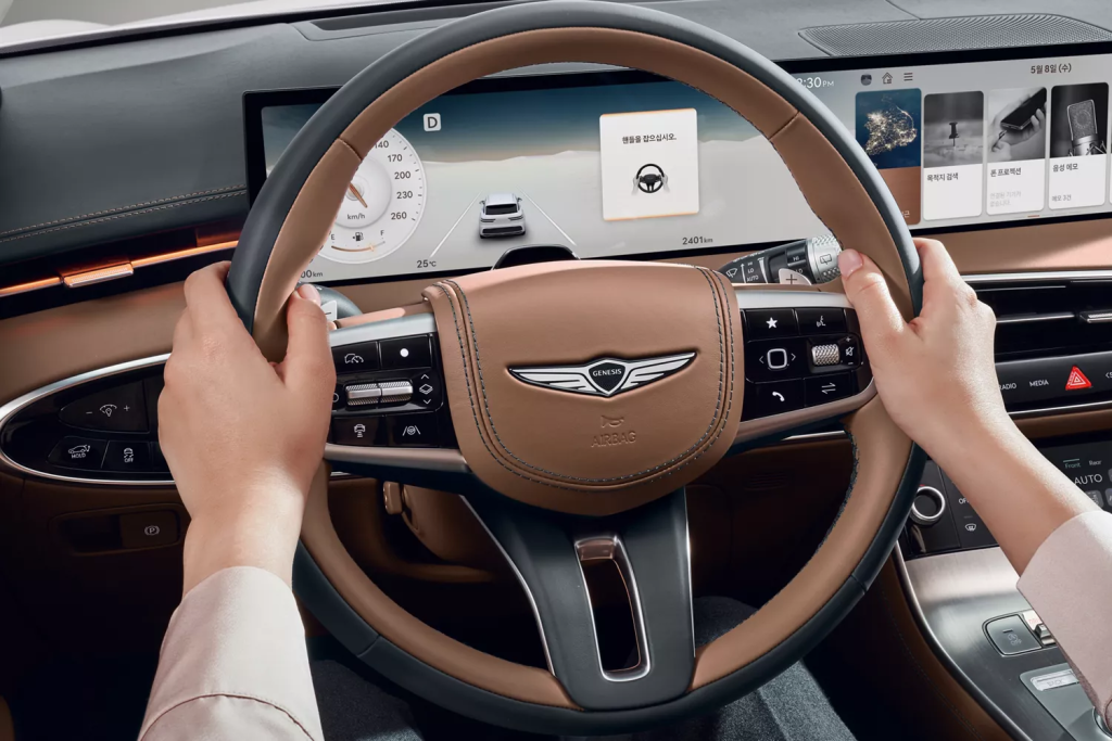 A Person holding a Genesis car steering wheel with a digital dashboard display showing driving information and controls.