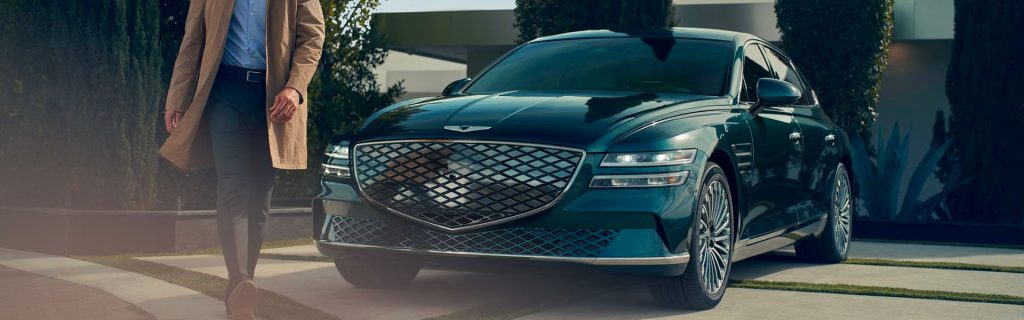 A person in a tan coat walks beside a dark green Genesis luxury sedan parked on a modern driveway with greenery in the background.