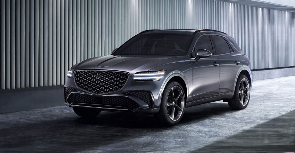 A silver Genesis GV70 SUV is parked indoors under artificial lights, featuring a large front grille and sleek design.