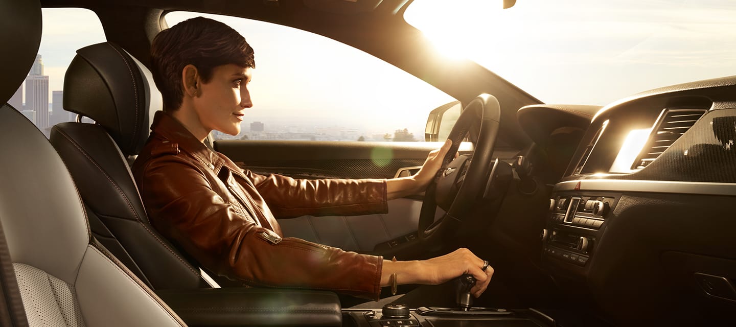 A person sitting in the driver's seat of a car, wearing a brown leather jacket, with sunlight illuminating the interior.