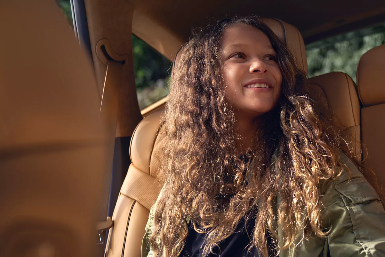 A young girl with curly hair is sitting in a Genesis GV70 car, smiling while looking out the window.