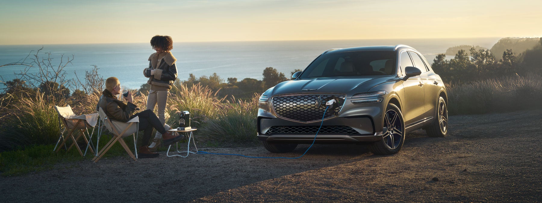 Silver Genesis electric SUV charging at a scenic coastal overlook, with two people relaxing nearby.
