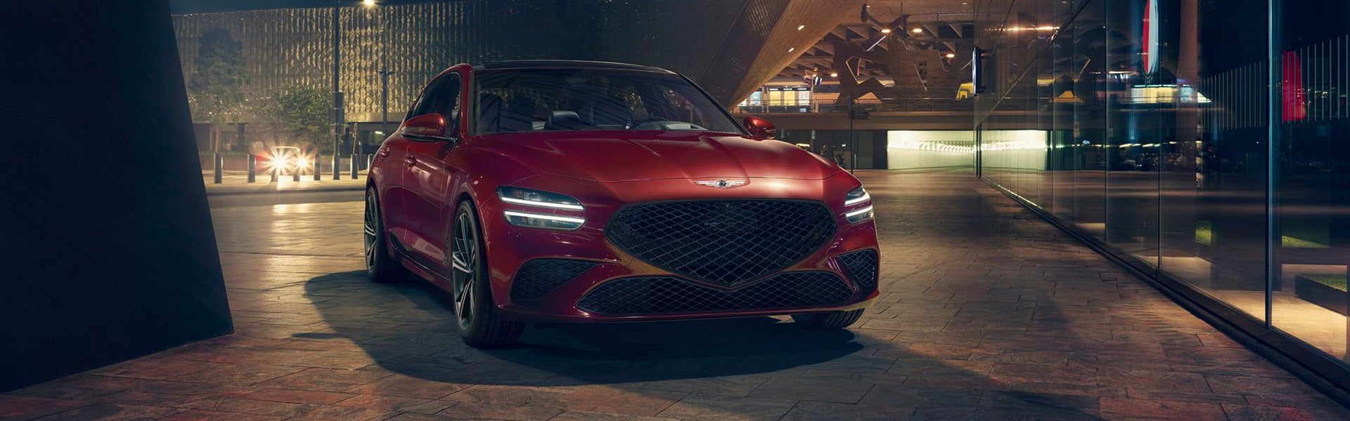 A sleek luxury sedan parked at dusk in a city setting, its front grille and headlights glowing under ambient street lighting.