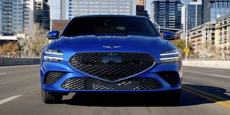 A blue Genesis G70 driving on a city road with modern buildings in the background.