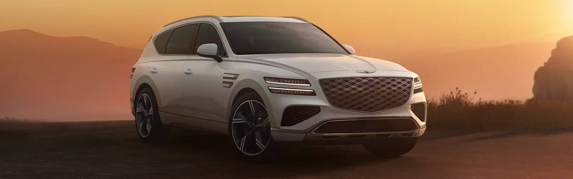A white Genesis GV80 with a sleek design parked on a dirt road during sunset, showcasing mountains in the background.