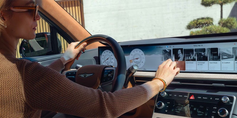 A woman driving a Genesis GV80, interacting with a digital dashboard display that shows various options and gauges.